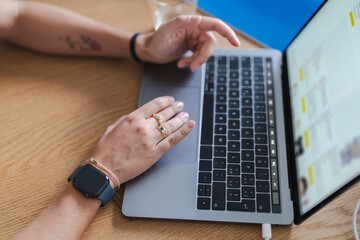 A person uses a laptop on a wooden table, browsing an online store. One hand rests on the trackpad, the other hovers above the keyboard. They wear rings and a smartwatch.