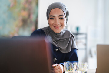 A happy young woman in a grey hijab smiles while working on her laptop. She is engaged in remote work or online learning from a bright home office. Empty glasses are on the desk.