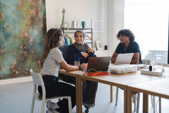 Three diverse young adults collaborate at a modern office table. Two women and one man work on laptops and discuss ideas in a bright, creative workspace.