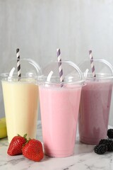 Milkshakes of different flavors in plastic cups and ingredients on white marble table, closeup