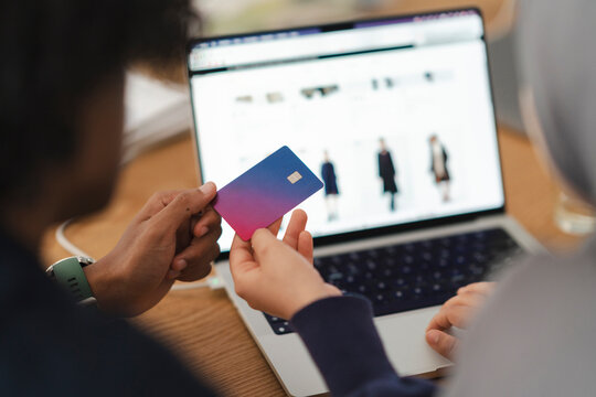 Two people are online shopping. One holds a card, ready to input payment details into a laptop displaying an e-commerce website with clothing. Digital transaction.