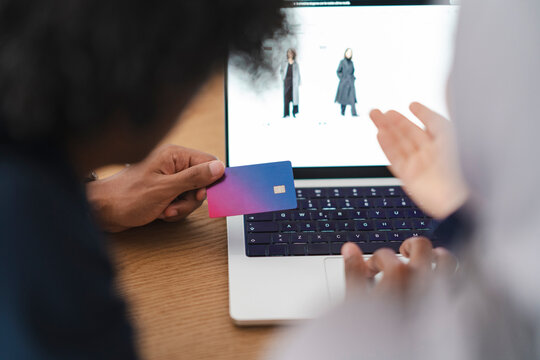 Two people engage in online shopping on a laptop. One holds a credit card, preparing for payment. The other navigates the e-commerce website, browsing clothing items.