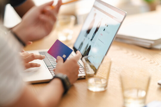 A person holds a credit card while shopping online on a laptop. They are browsing an e-commerce website, preparing to make a purchase. Another person's hand is visible.