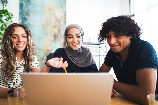 Three diverse young adults, two women and one man, are collaborating around a laptop. One woman points at the screen with a pencil, all smiling.