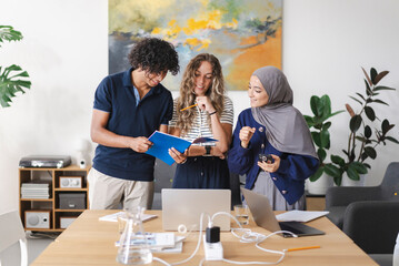 A diverse group of three young adults, including a man and two women, one wearing a hijab, collaborate around a table. They are smiling, discussing content in a blue notebook