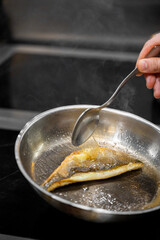 Chef basting seared fish fillet in hot oil using spoon in stainless steel pan. Steam rises, skin crispy. Professional cooking technique in action