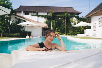 Confident woman touching hair while leaning on pool edge, embracing digital lifestyle, clarity of purpose and relaxed luxury made possible by remote working technologies.