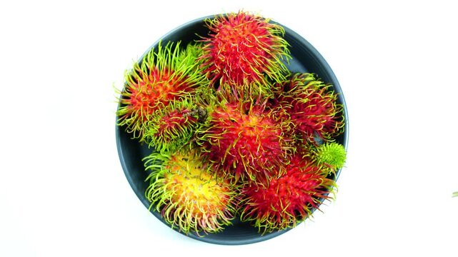 Top view rotation of ripe sweet rabutan fruits in black ceramic dish on white background.