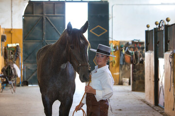 A young, attractive woman dressed in riding clothes and a traditional Spanish hat is standing in...