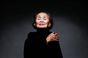 Elderly woman portrait body language serene expression in studio lighting with closed eyes and hand on shoulder wearing dark sweater