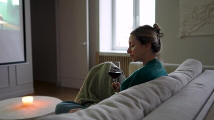 Cozy evening of woman sitting on sofa with blanket eating snack, drinking wine, watching movie on home projector. Relaxed female with glass of red wine enjoys cinematic mood, comfort with candlelight  - Powered by Adobe