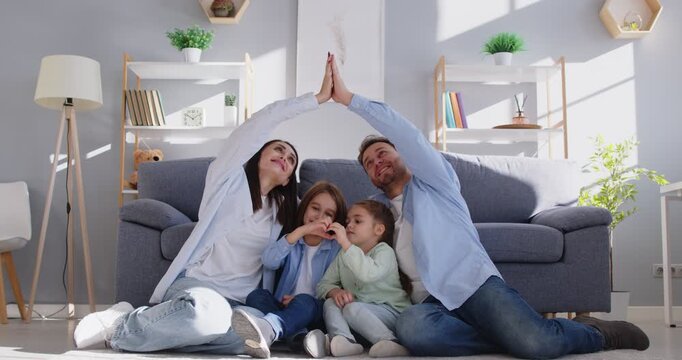 Happy family care at new home, young mother and father with two little children sitting on floor making hand house roof, tender moment, smiling parents protect children, keep safe from harm or injury