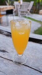 Refreshing orange iced beverage served in a tall glass, adorned with ice cubes, placed on a weathered wooden table in a blurred, tranquil outdoor garden or cafe setting