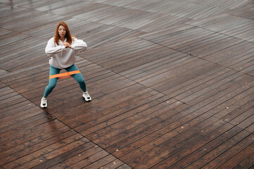 Obraz premium Sporty fit woman with red hair squats with fitness elastic band on wooden pier