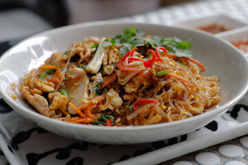 Close up A white bowl of stir-fried glass noodles with vegetables and herbs, served alongside fresh spring rolls and dipping sauces on a patterned table.