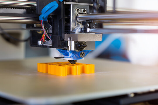 Close-up of a desktop three-dimensional printer nozzle depositing molten filament to build a yellow plastic prototype on the heated bed, showcasing additive manufacturing process and precision.