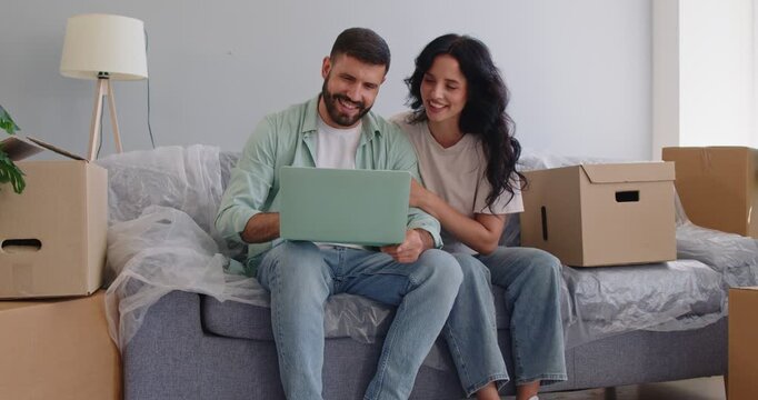 Happy couple using laptop among moving boxes in new home, young people sitting on sofa in living room. Young smiling man and woman plan relocation tasks and schedule of delivery, future renovation