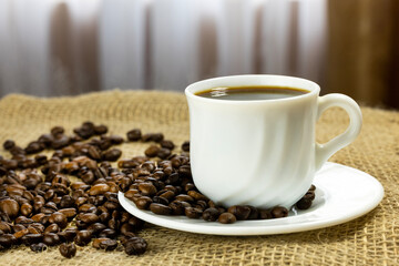 A white cup filled with black coffee sits on a saucer. Coffee beans are scattered around the cup on a burlap surface. Natural light filters through a window.
