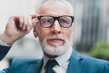 Confident mature businessman wearing glasses in formal blue suit standing outdoors with focus and...