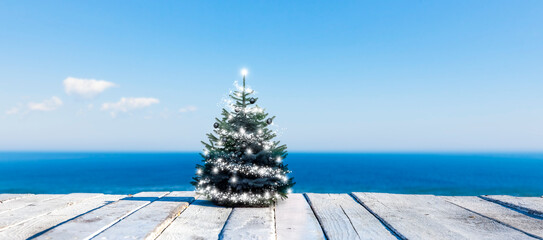 Geschmückter Weihnachtsbaum auf einem weissen Holzdeck vor einem Blauen Meer