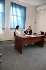Two women in business suits sit at a desk, talking and drinking water during a business meeting.