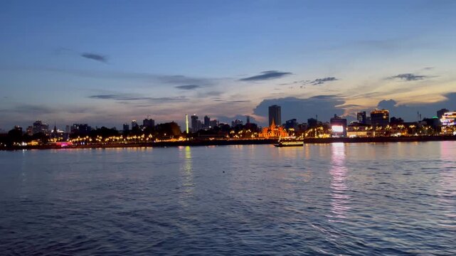 Sunset view of the Phnom Penh riverfront in Cambodia, featuring the illuminated Royal Palace boat sailing and surrounding skyline. Cultural heritage with modern high-rises blending concept