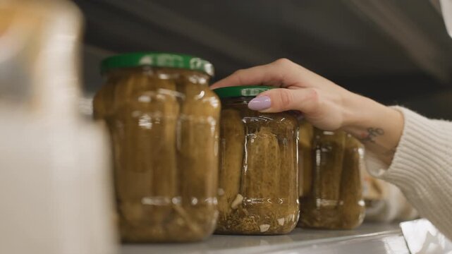 Close up of mall goer hand returning glass jar of pickles with green lid to shelf in supermarket aisle with blurred background highlighting nails metal rails and retail during shopper browsing