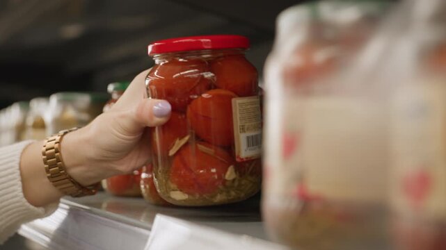 Close up of female customer hands adjusting transparent glass jar of tomatoes on metal shelf rails in supermarket aisle highlighting polished nails wrist watch blurred bright busy grocery background