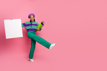 Cheerful young woman holding a blank poster and yoga mat against a vibrant pink background showcasing casual fashion.