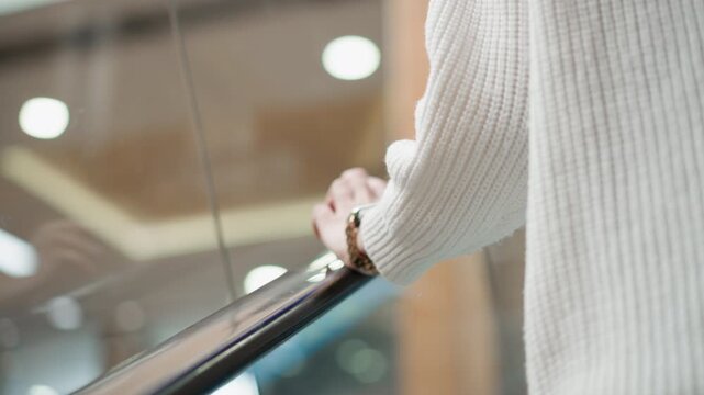 close up of demure woman wearing wrist watch tapping handle of moving walkway in modern mall corridor under bright overhead lights beside glass railing capturing gentle motion and casual elegance