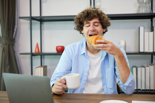 young man eating croissant while working on laptop computer at home - Powered by Adobe