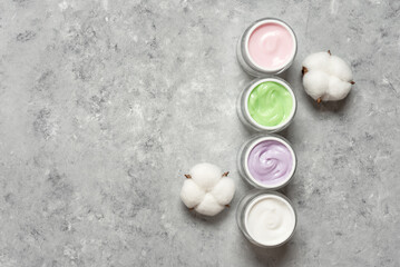 Colored cosmetic creams in jars with cotton flowers on grey concrete background, top view, copy space