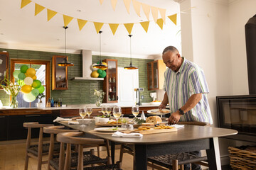 Senior Asian man arranging cheese board and wine glasses on dining table in kitchen, copy space
