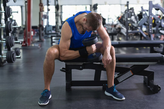 Man sitting on weight bench performing concentration curls in gym wearing sportswear with dumbbell - Powered by Adobe