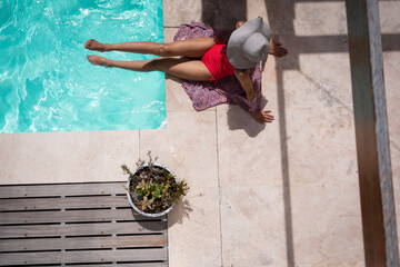 Woman sitting beside rectangular pool on sarong on patio wearing sun hat near planter and bench