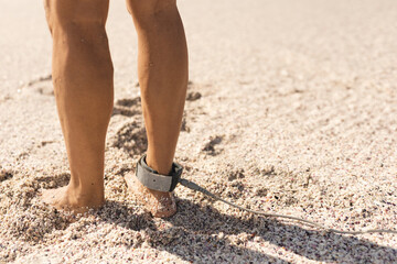Man standing barefoot on coarse beach sand near shoreline, wearing neoprene surfboard leash cuff