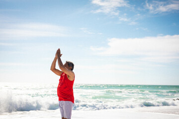 Senior man in red shirt stretching arms overhead at sandy shoreline with ocean waves, copy space