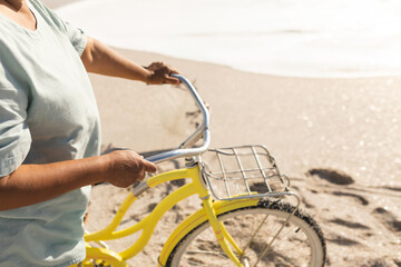 Senior woman holding yellow bicycle with front rack, standing on sandy beach near ocean, copy space