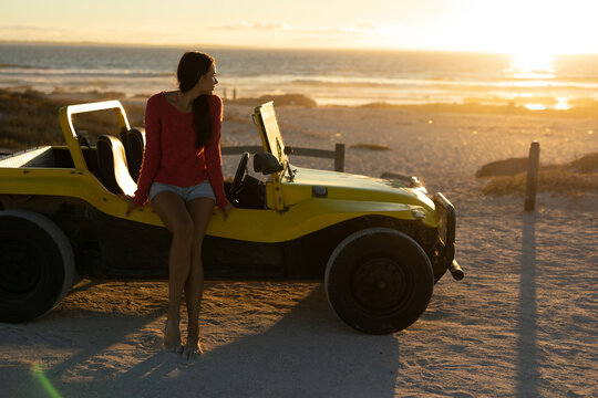 Woman sitting on yellow dune buggy at beach at sunset, gazing ocean and posts, copy space - Powered by Adobe