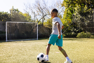 African American boy wearing white tee and shorts dribbling soccer ball on turf pitch, copy space
