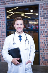 Doctor standing in front of brick wall and glass window, holding black folder and stethoscope