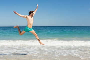 Man wearing turquoise swim shorts and dark sunglasses leaping above surf on sandy beach, copy space
