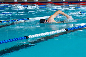 Male swimmer performing front crawl at aquatic center pool with swim cap, goggles and lane ropes
