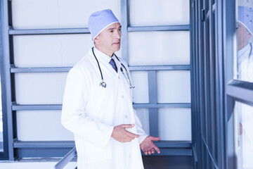 Doctor is standing beside glass panel in hospital corridor with stethoscope and surgical cap