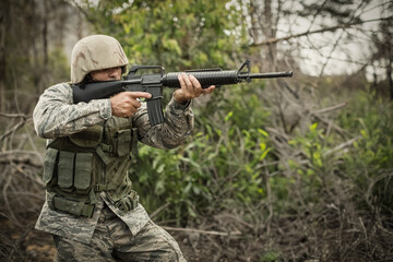 Male soldier kneeling, aiming rifle and wearing vest and camouflage helmet in forest, copy space