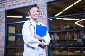 Doctor standing holding stethoscope and blue clipboard in medical corridor revealing bookshelves