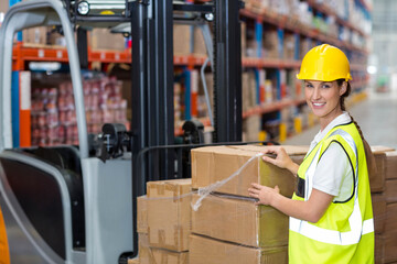 Warehouse worker standing beside palleted boxes, wearing vest and hard hat near forklift and racks