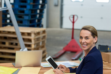 Woman holding and using tablet at warehouse desk with laptop, boxes, pallet jack and pallets