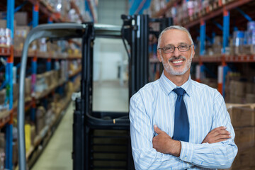 Supervisor standing in bright warehouse near idling forklift and racks stocking boxes and pallets