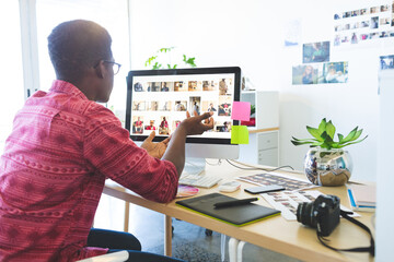African American man sitting at creative studio desk reviewing thumbnails on computer using tablet
